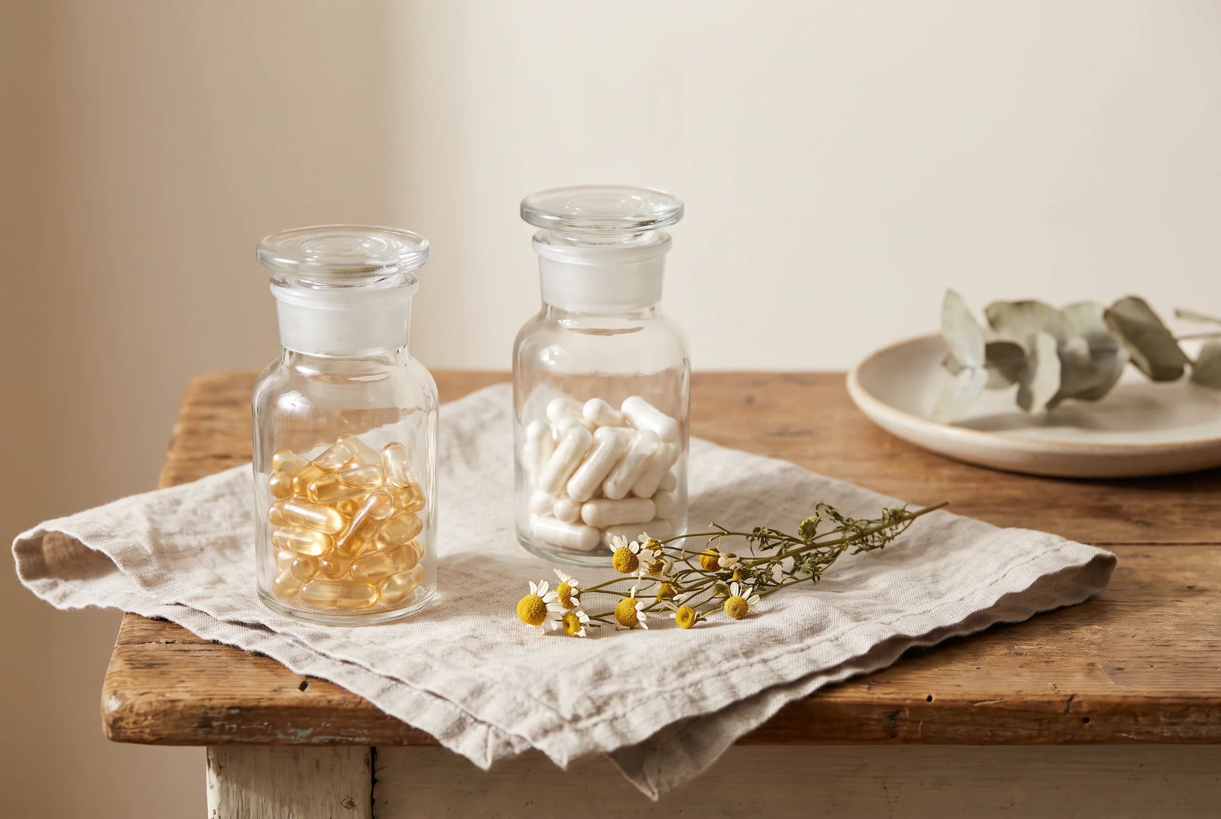 Two unlabeled clear glass apothecary jars resting on a soft linen napkin, one with golden capsules and one with white powder capsules, separated by a sprig of dried chamomile.